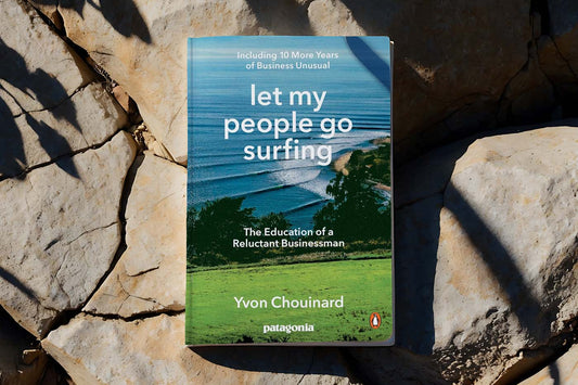 Cover of 'Let My People Go Surfing' by Yvon Chouinard. The background shows a coastal scene with green fields in the foreground and blue ocean waves rolling toward the shore.