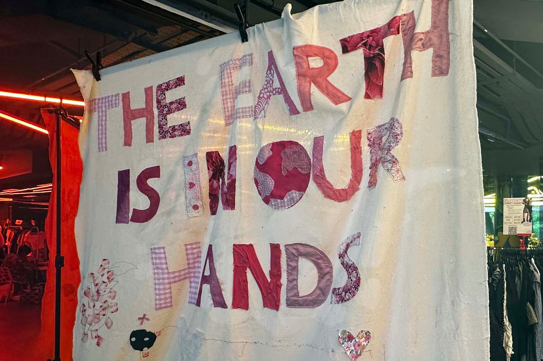 A handmade fabric banner with patchwork letters reading "THE EARTH IS IN OUR HANDS," displayed at a sustainable fashion event, surrounded by upcycled clothing.